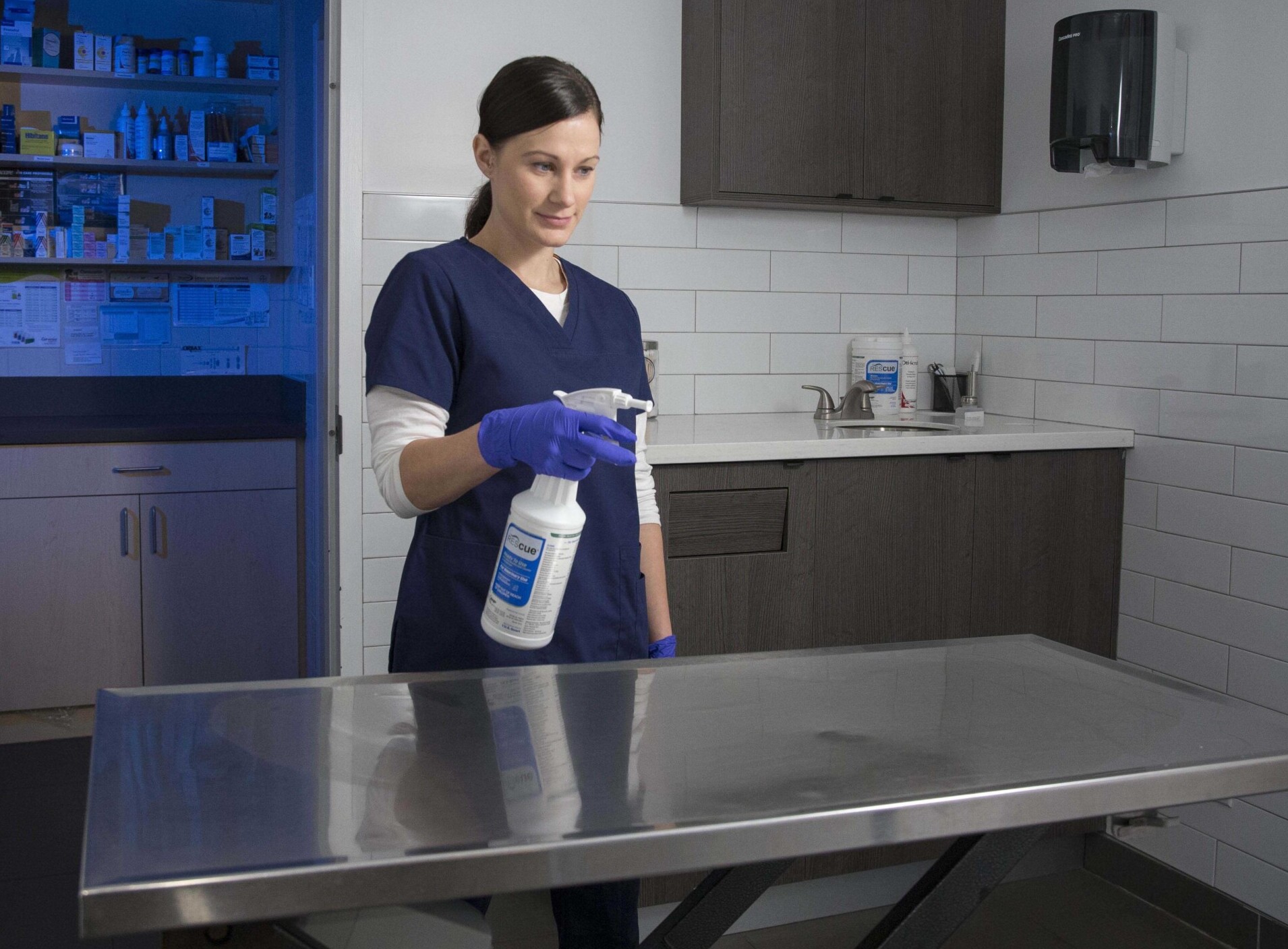 Veterinary professional wearing gloves applying Rescue disinfectant spray to a stainless steel exam table.